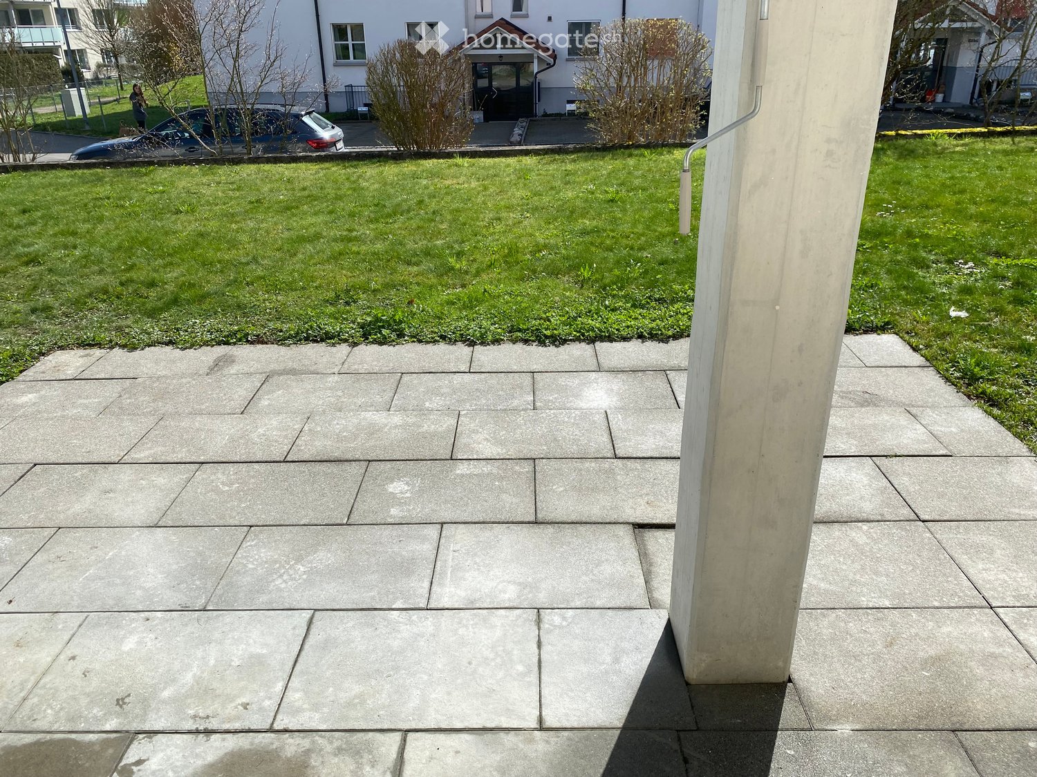 Concrete patio with a grassy lawn, small bushes, and a building entrance in the background.