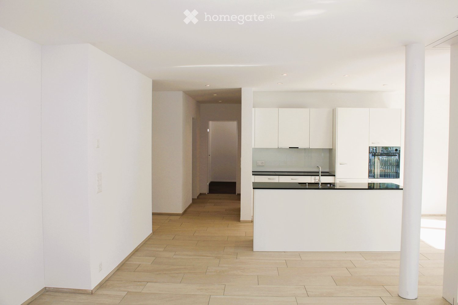 Empty room with light-colored walls, wooden flooring, and a modern kitchen area visible in the background. No furniture or personal items are present.