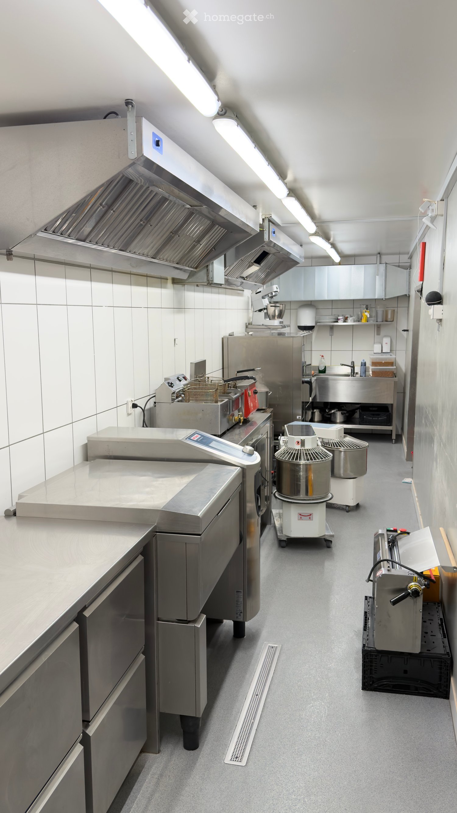 Commercial kitchen with stainless steel appliances, including a refrigerator, mixer, oven, fryer, and sink, arranged around a central counter.