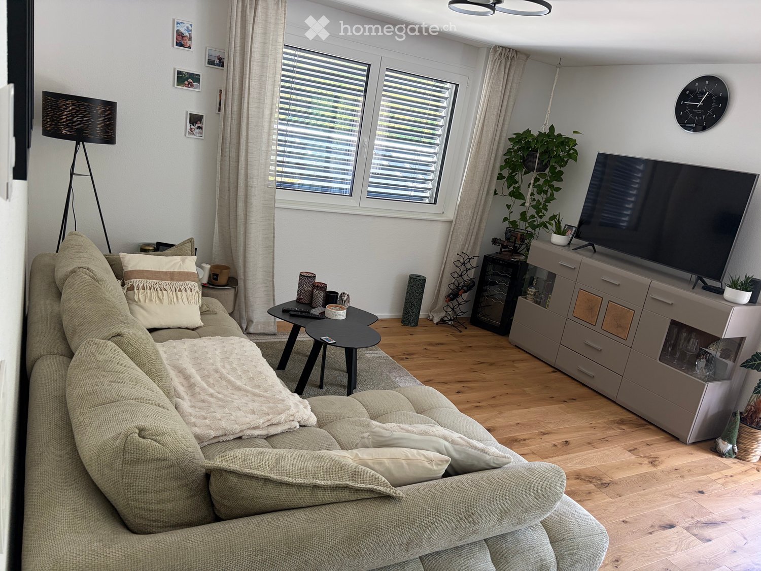 Modern living room with a beige sofa, coffee table, television, side table, floor lamp, clock, and decorative plants.