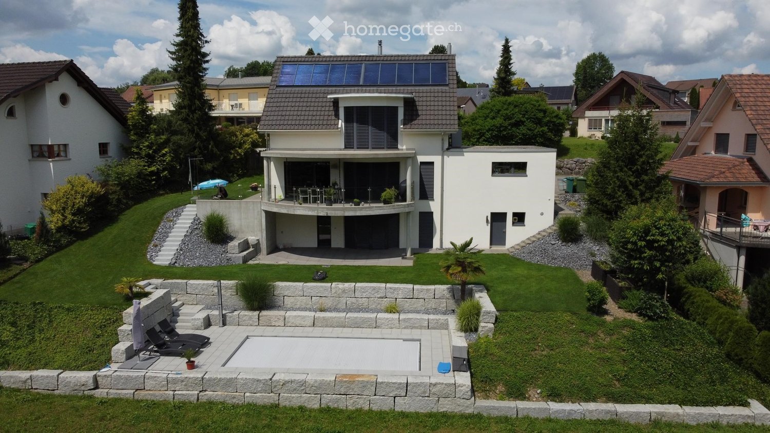 A modern two-story house with a large balcony, surrounded by a well-maintained garden with a swimming pool, lounge chairs, and various plants.