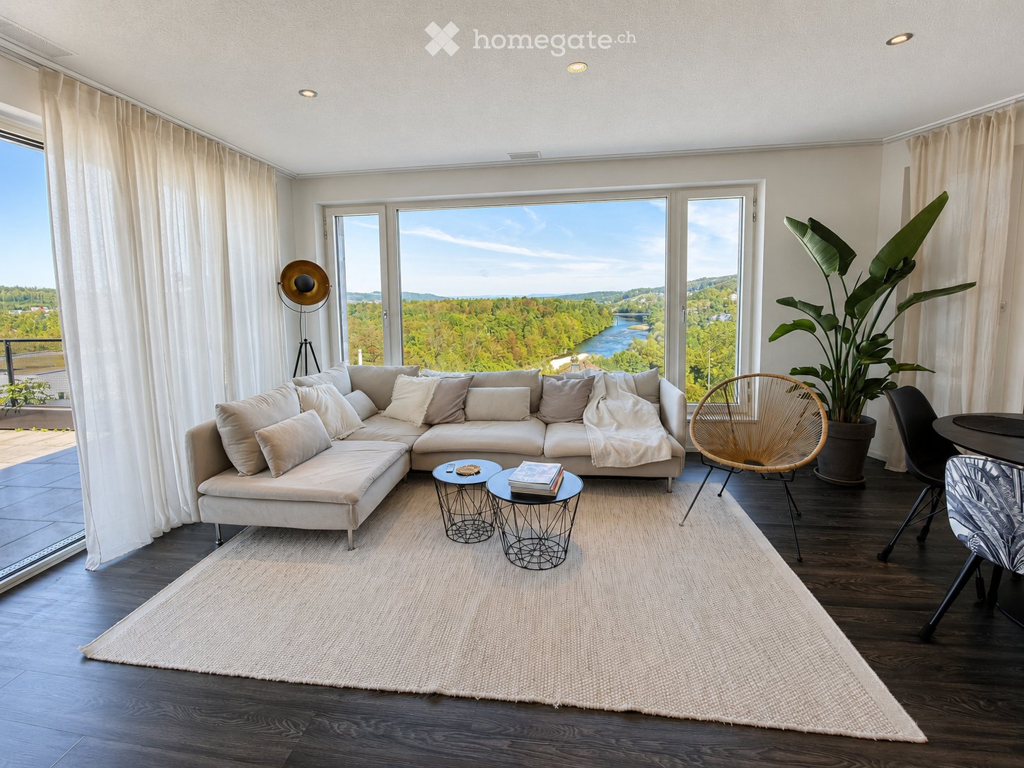 Modern living room with large windows offering a scenic view, featuring a sectional sofa, coffee tables, a wicker chair, and a large plant.