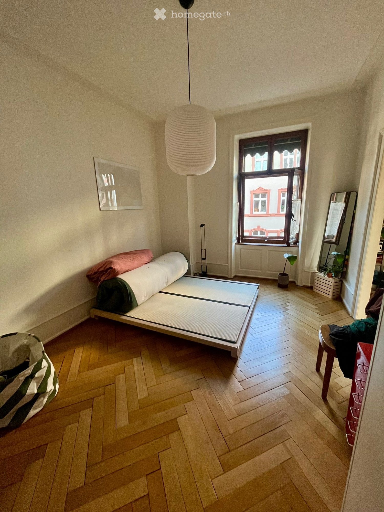 Minimalist bedroom with tatami mat bed, wooden floor, large window, hanging lamp, small stool, mirror, and potted plant.