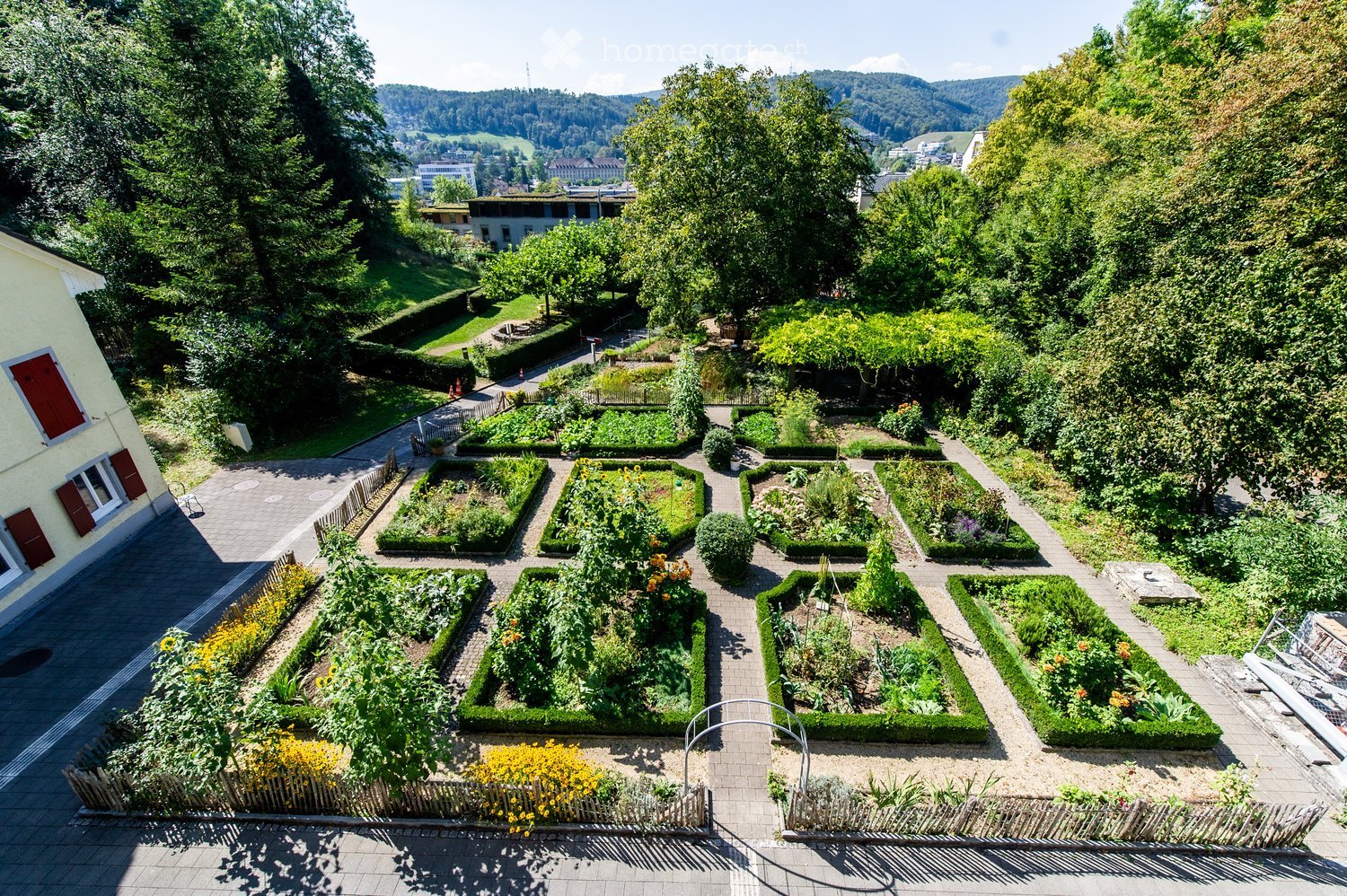 A well-maintained garden with neatly arranged flower beds, pathways, and surrounding greenery, featuring a variety of plants and shrubs.