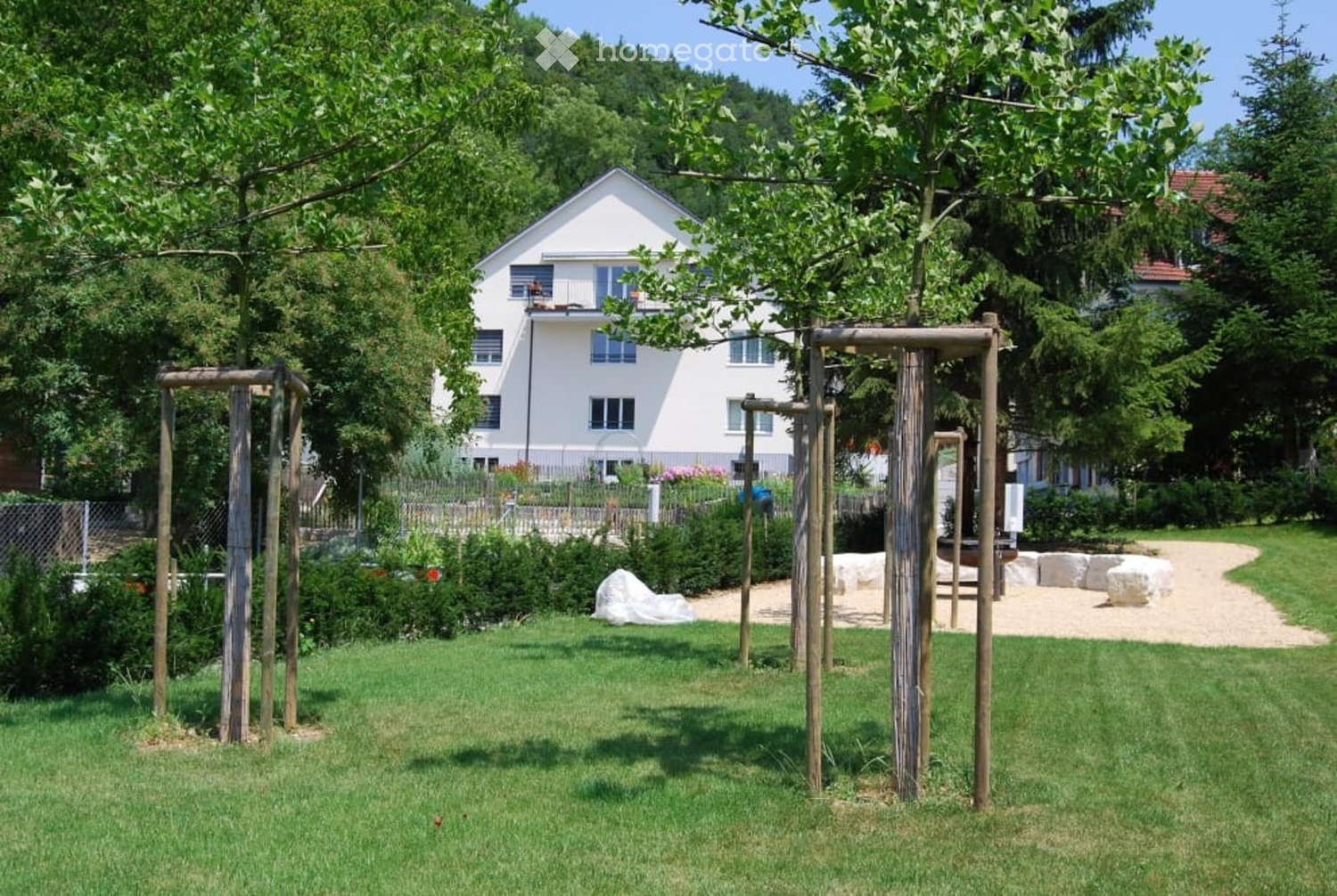 green lawn, wooden structures, trees, white house in the background, gravel path