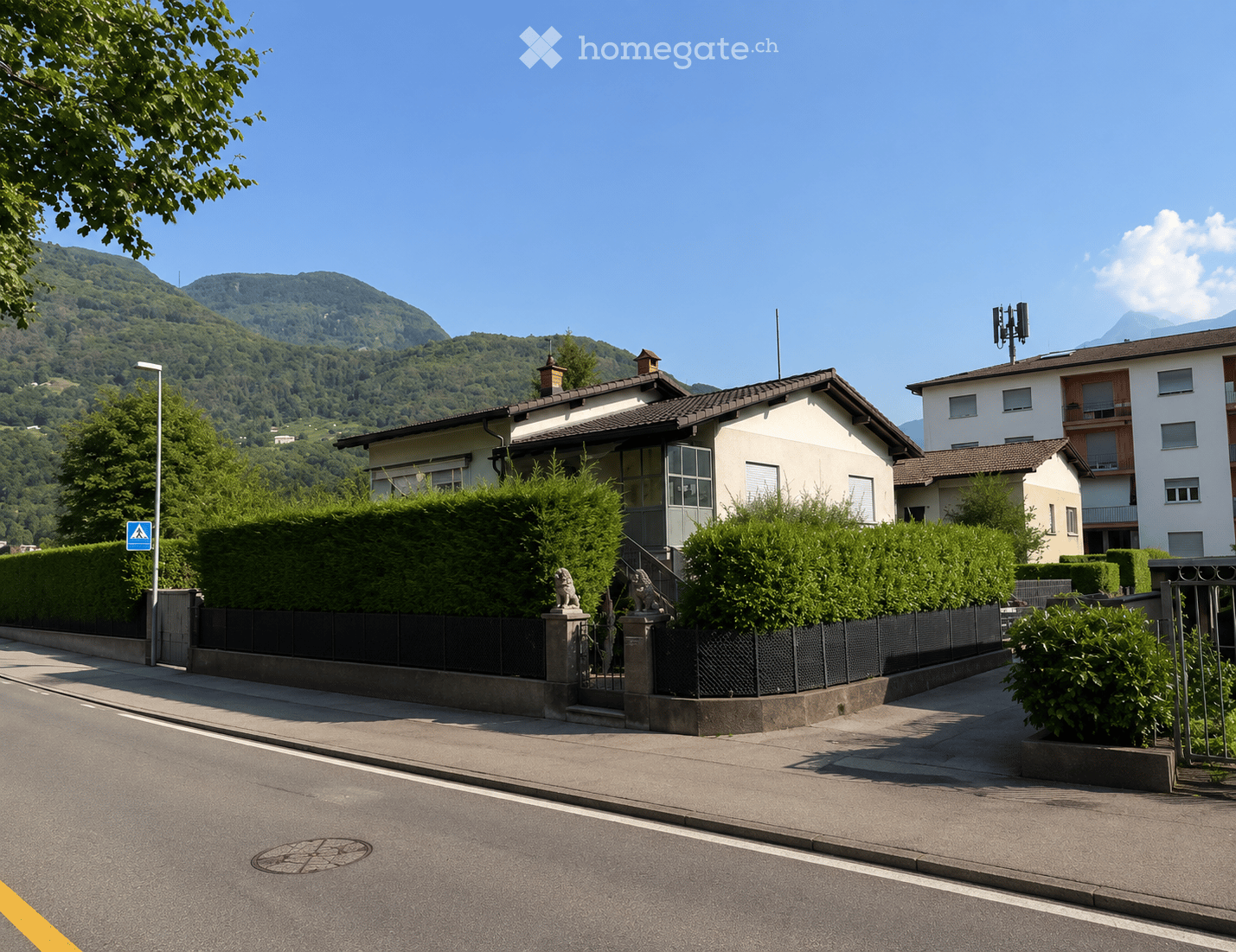 A two-story house with a tiled roof, surrounded by greenery and a black metal fence, with a view of mountains in the background.