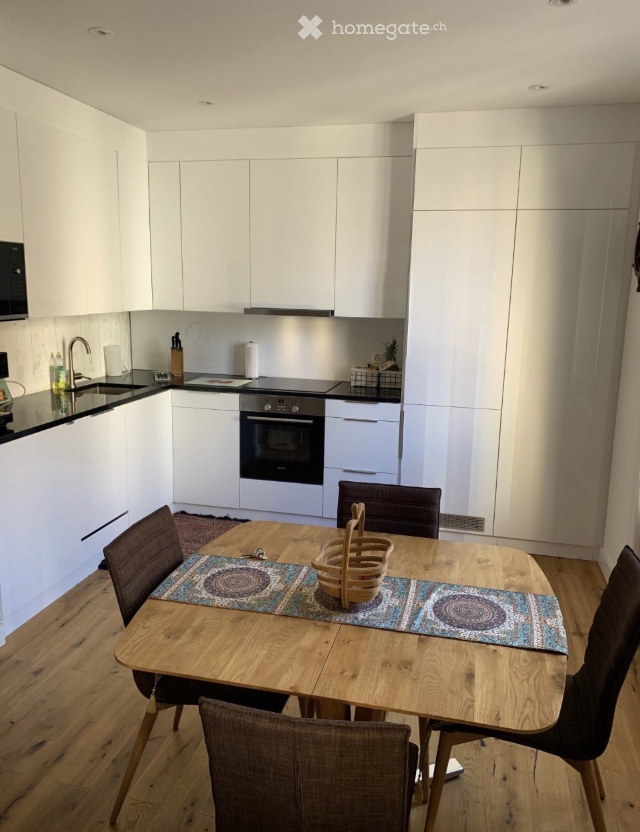 Modern kitchen with white cabinets, a wooden dining table with four chairs, and a small basket on the table. The kitchen features an oven, microwave, and sink.