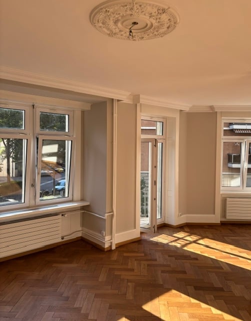 Empty room with wooden flooring, large windows, and a decorative ceiling light fixture. No furniture or equipment present.