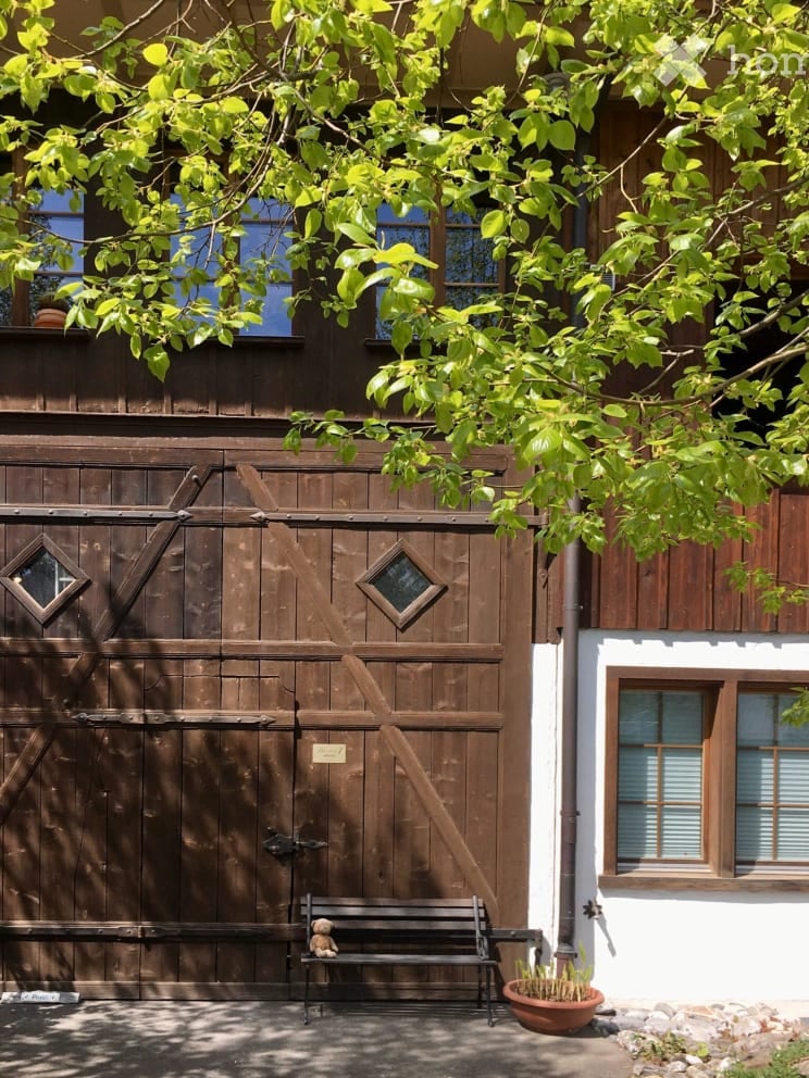 A house with wooden exterior, white window frames, a wooden garage door with diamond-shaped glass windows, a bench with a teddy bear, and a potted plant