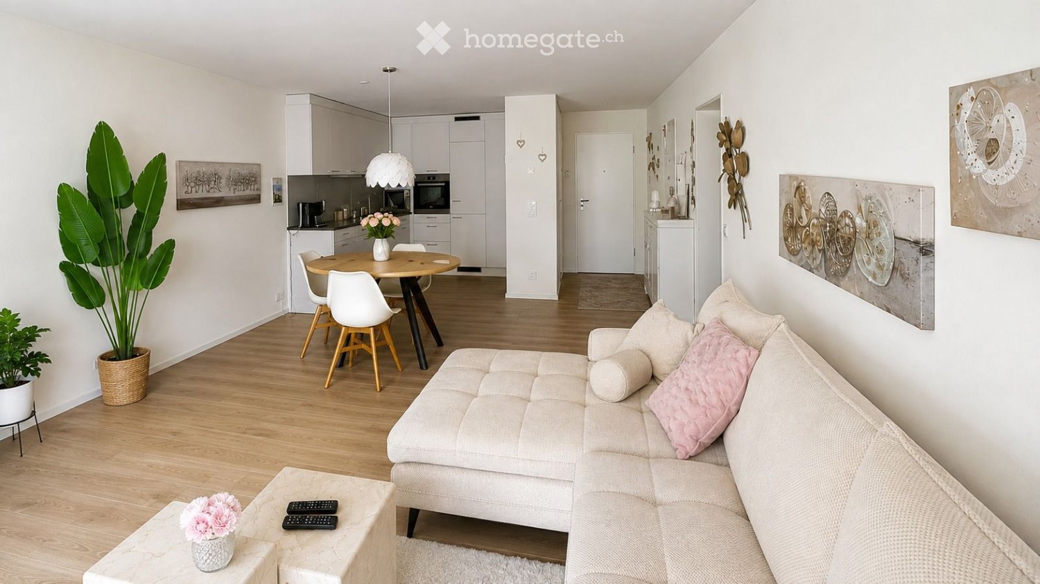 Modern living and dining area with light wood flooring, white walls, a beige sectional sofa with pink cushions, a small coffee table with remote controls and a vase of flowers, a dining table with four chairs, a kitchen area with white cabinets and applia