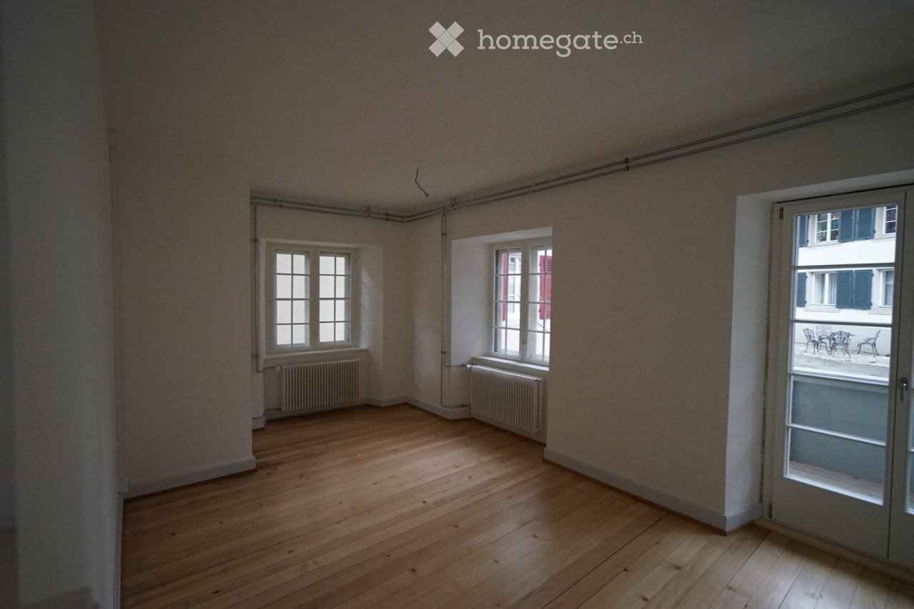 Empty room with wooden flooring, white walls, and multiple windows, including a sliding glass door leading to a balcony area.