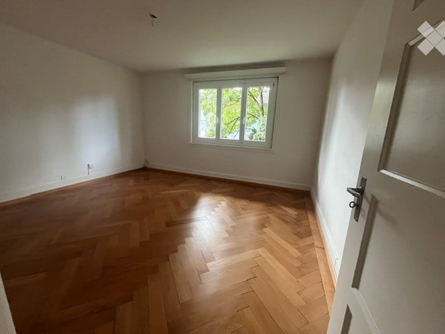 Empty room with wooden parquet flooring, white walls, and a single window letting in natural light. No furniture or equipment present.