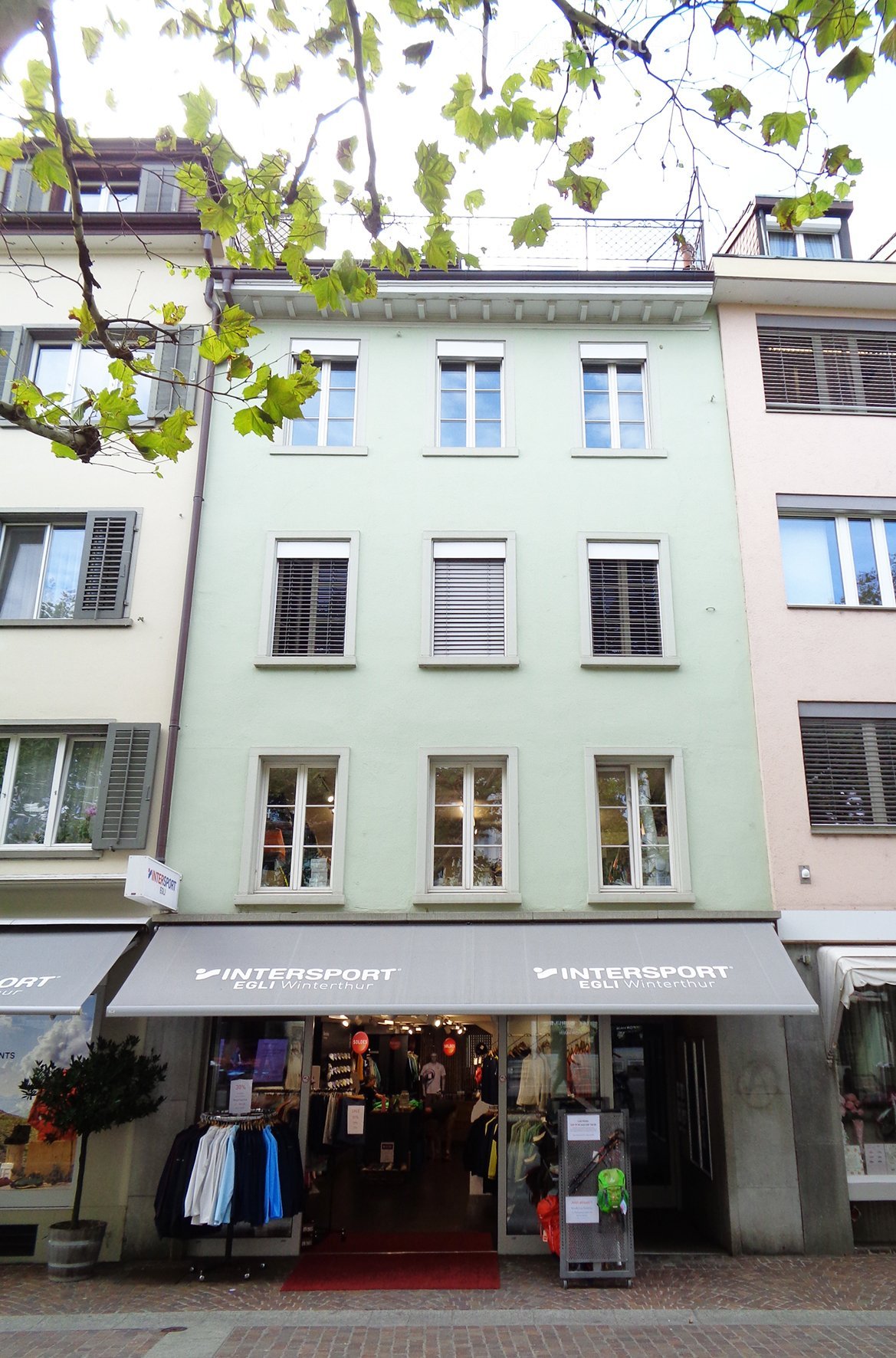 Three-story building with light green facade, multiple windows, and a storefront labeled "INTERSPORT EGLI Winterthur" with clothing racks and displays visible inside.