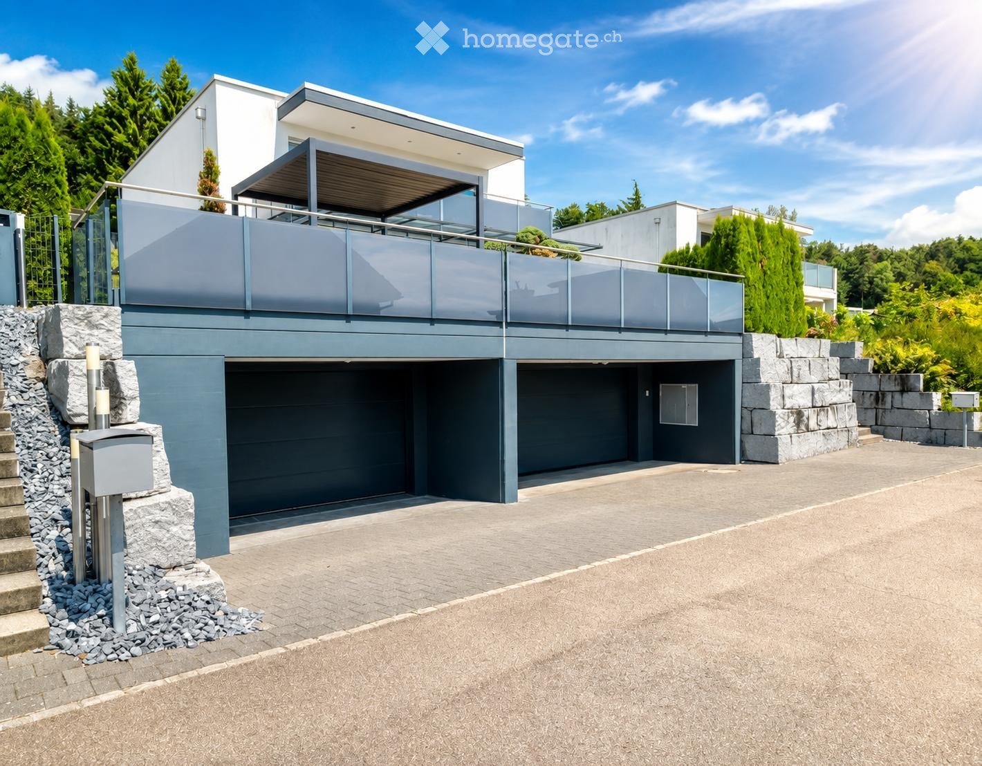 Modern two-story house with a grey garage, a raised balcony, and a stone staircase leading to the entrance.