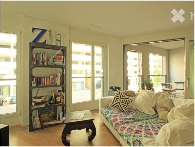 Bright living room with large windows offering a city view, featuring a colorful patterned sofa, wooden coffee table, bookshelf filled with books, and a small dining area with a table and chairs.