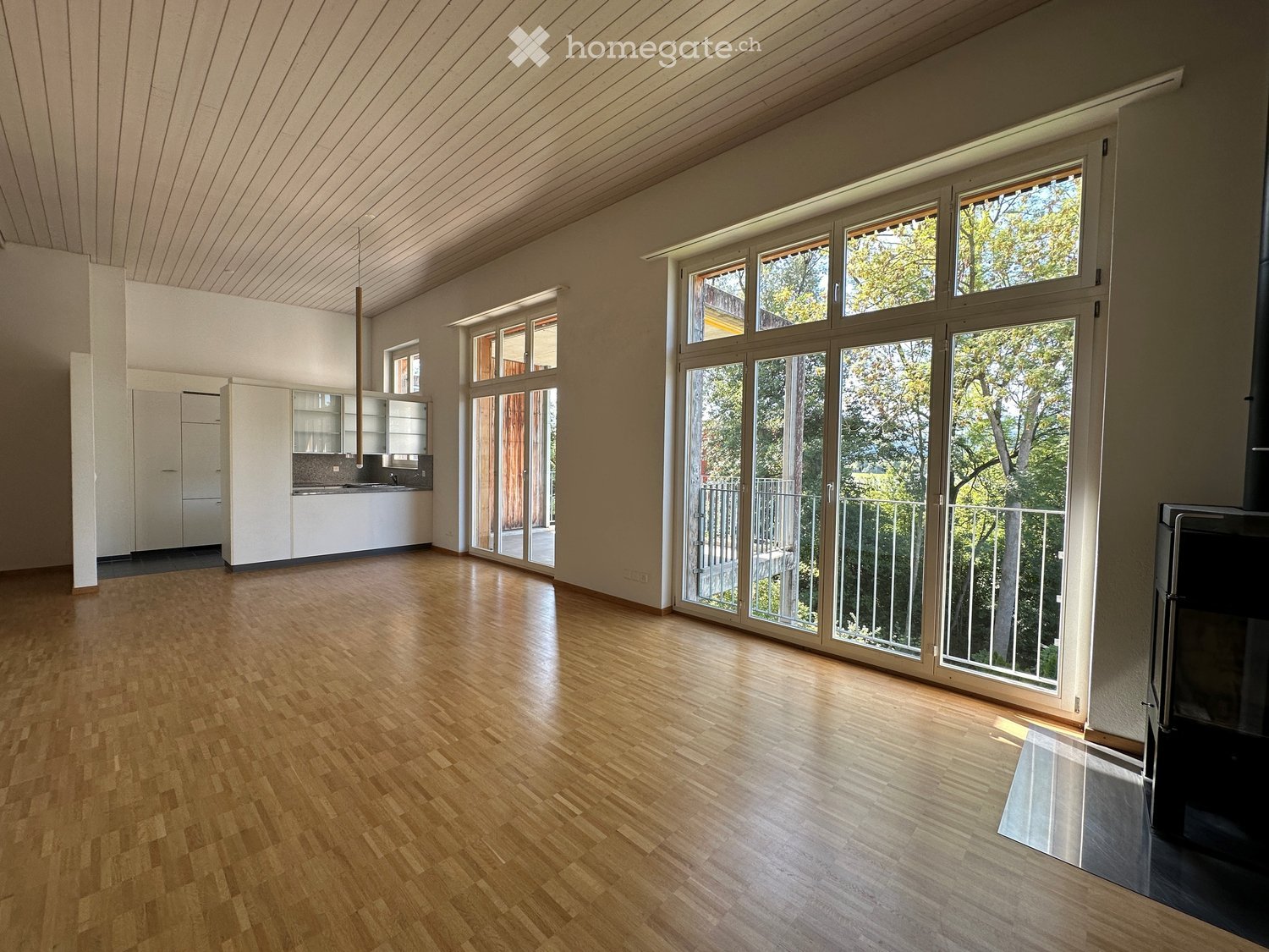 Bright, open-plan room with wooden flooring, large windows and sliding doors leading to a balcony, and a modern kitchen area with white cabinets and stainless steel appliances.
