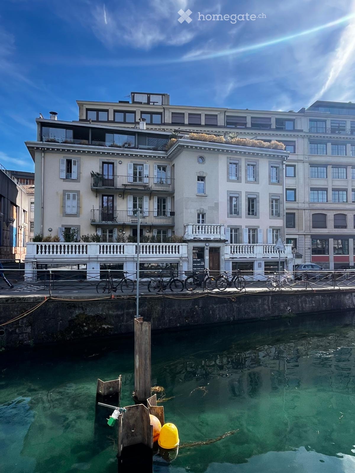 A multi-story building with balconies and large windows, situated near a waterfront with bicycles parked along the pier.