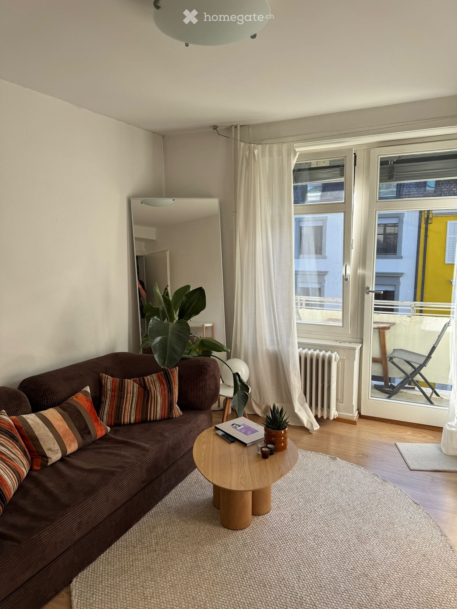 Modern living room with a brown sofa, wooden coffee table, large mirror, potted plants, and a view through large windows.