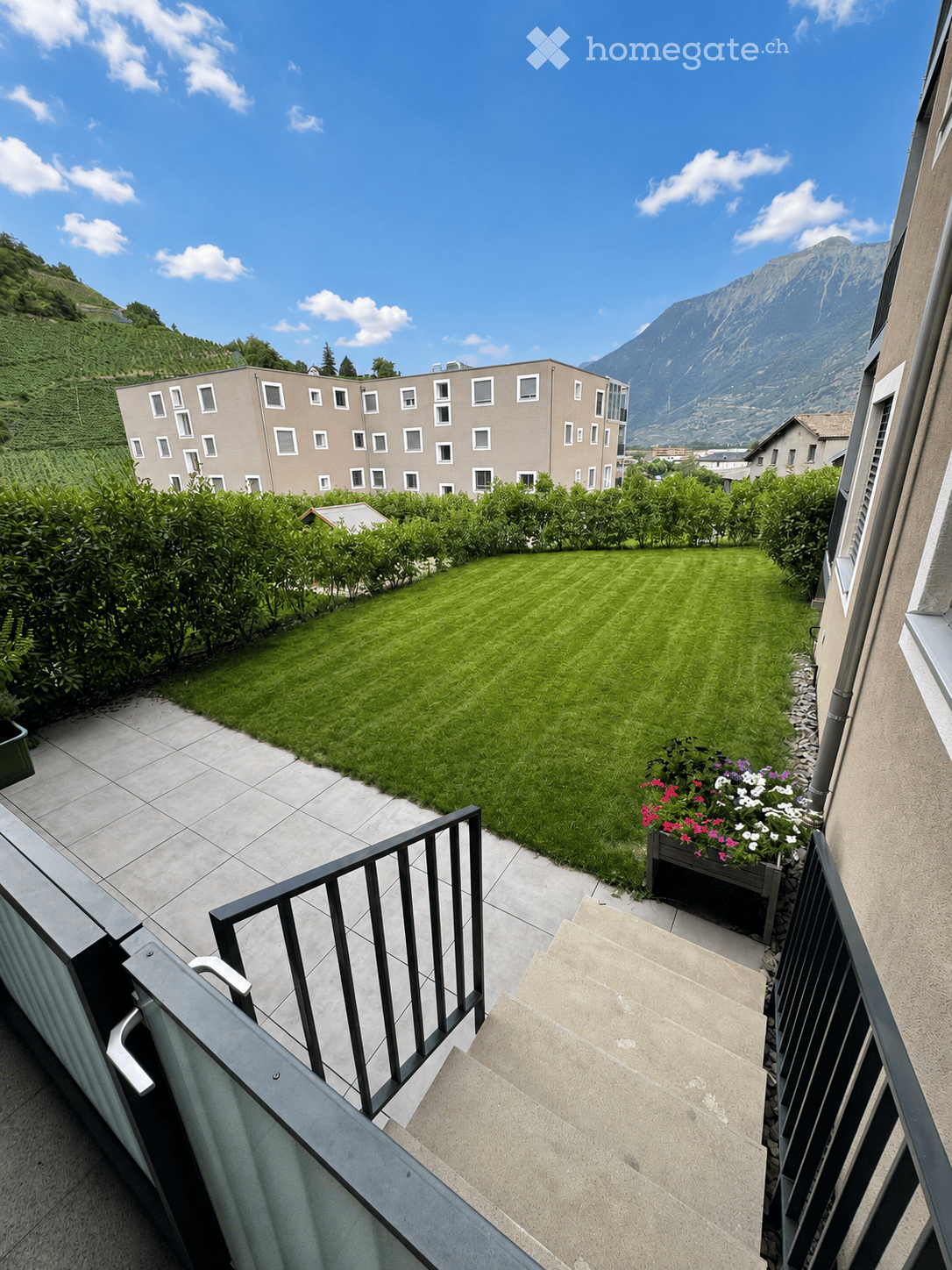 Well-maintained garden with a lawn, flower bed, and surrounding greenery, with a modern building and scenic mountain view in the background.