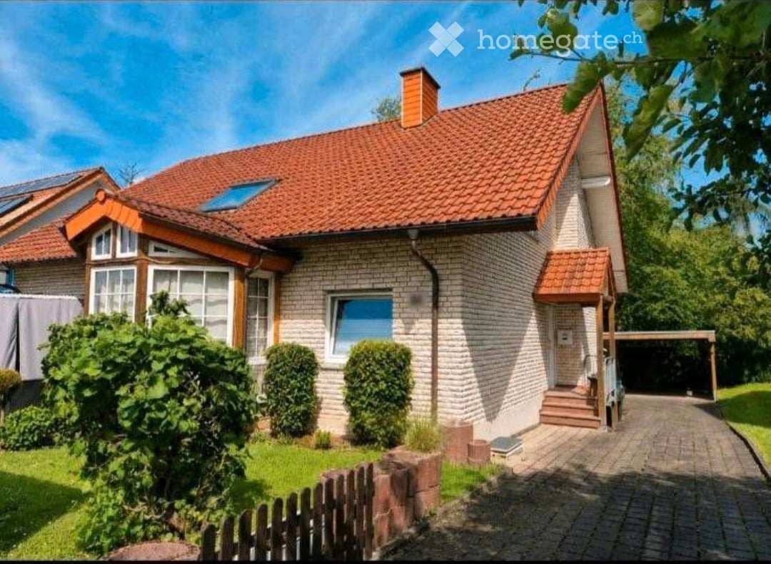 Two-story house with red-tiled roof, brick facade, and a small garage. The house features multiple windows, a chimney, and a well-maintained garden with a wooden fence.