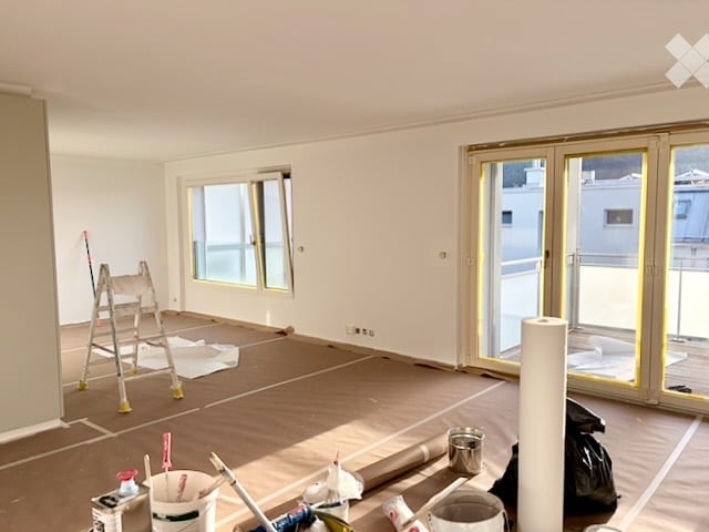 Empty room with white walls, large windows, and a sliding glass door leading to a balcony. Construction materials and tools are visible, indicating ongoing renovation.