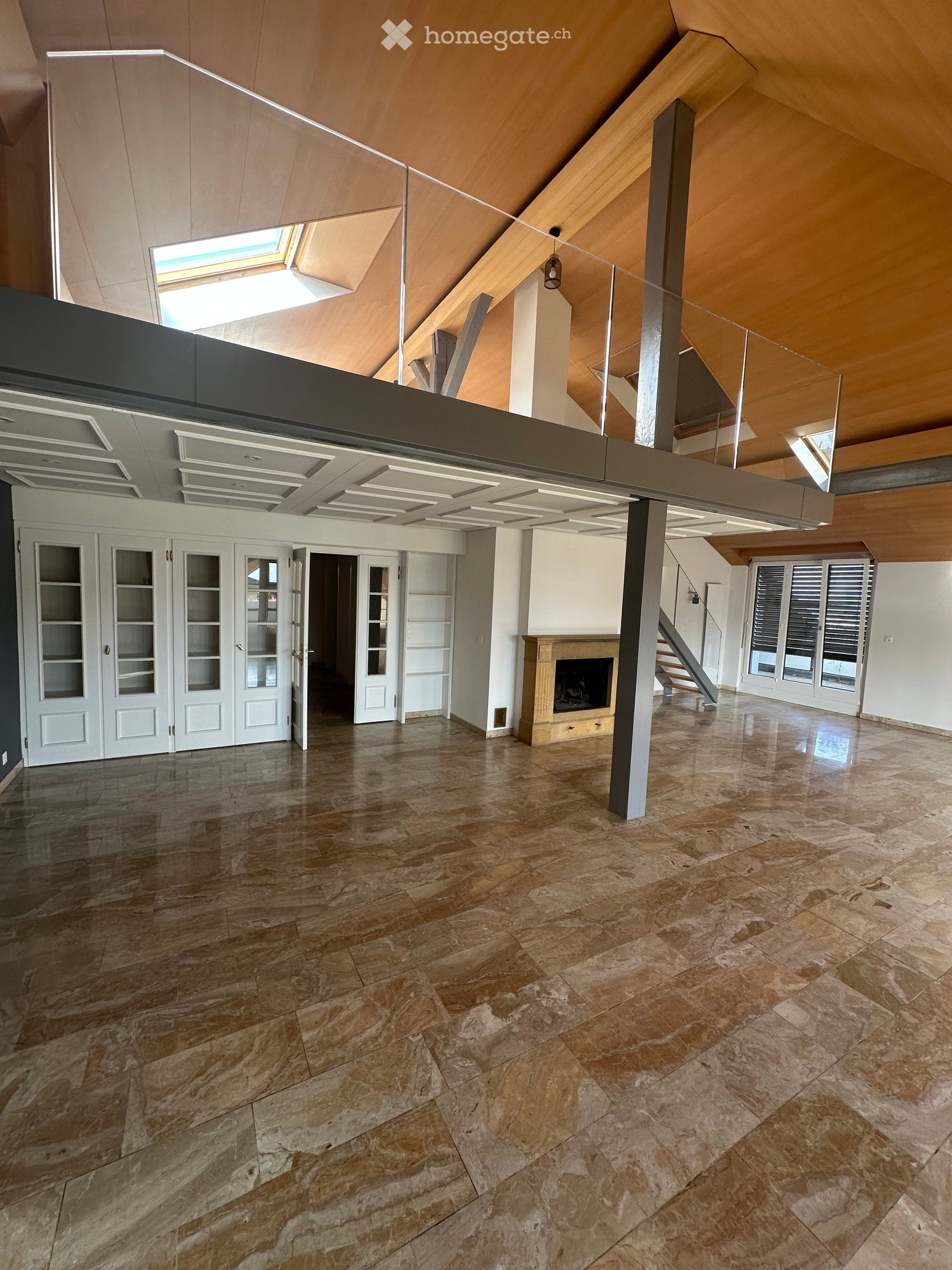 Spacious open-concept living area with high ceilings, wooden beams, and large windows. The room features a fireplace, built-in cabinets and shelves, and a tiled floor with a natural stone pattern.