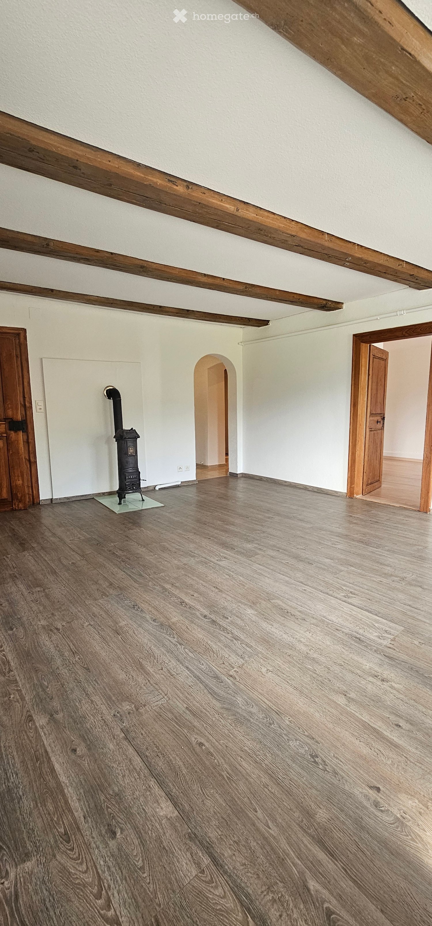 Empty room with wooden floor, white walls, exposed wooden beams, and a small black stove with a chimney. There are two wooden doors and an arched doorway leading to another room.