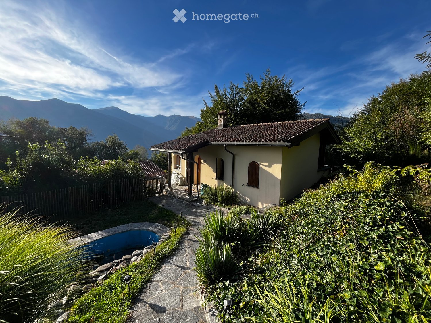 A small, single-story house with a tiled roof and light yellow walls, surrounded by a well-maintained garden with a swimming pool and lush greenery, offering a scenic mountain view in the background.
