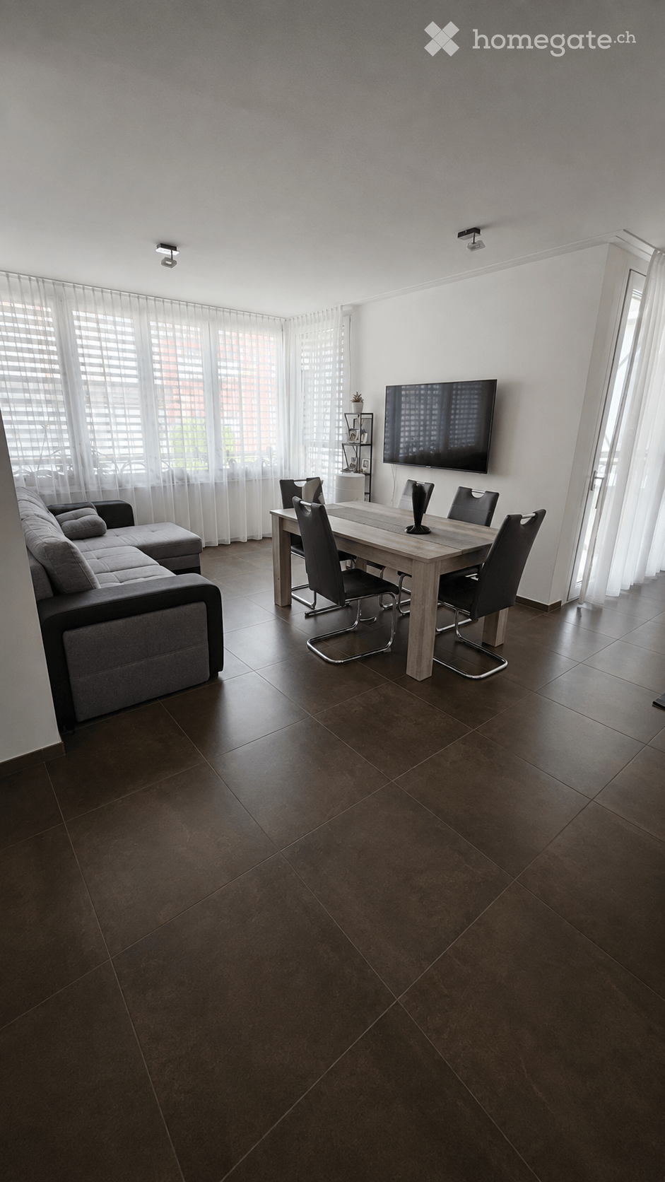 Modern living and dining area with dark brown tiled flooring, large windows with sheer curtains, a grey sectional sofa with an ottoman, a wooden dining table with four black chairs, a flat-screen TV mounted on the wall, and a small plant on a shelf.