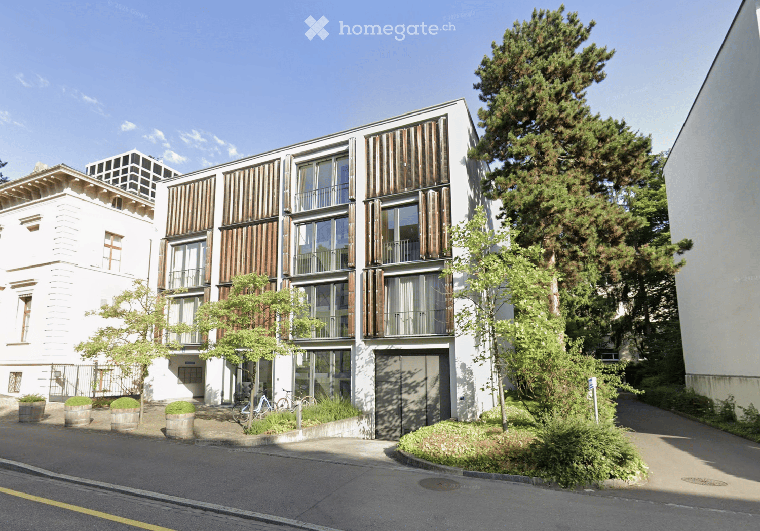Modern multi-story building with large windows, balconies, and contemporary design. Features include a mix of white and dark wood facades, greenery, and bicycles parked near the entrance.
