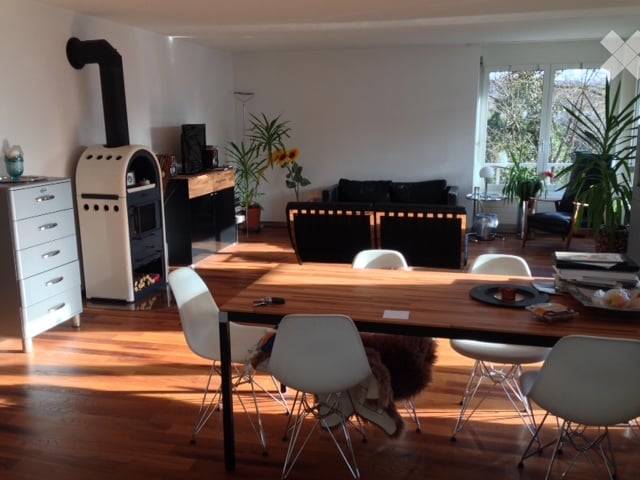 Bright living and dining area with wooden flooring, a large wooden dining table with white chairs, a black sofa, a fireplace, and potted plants.