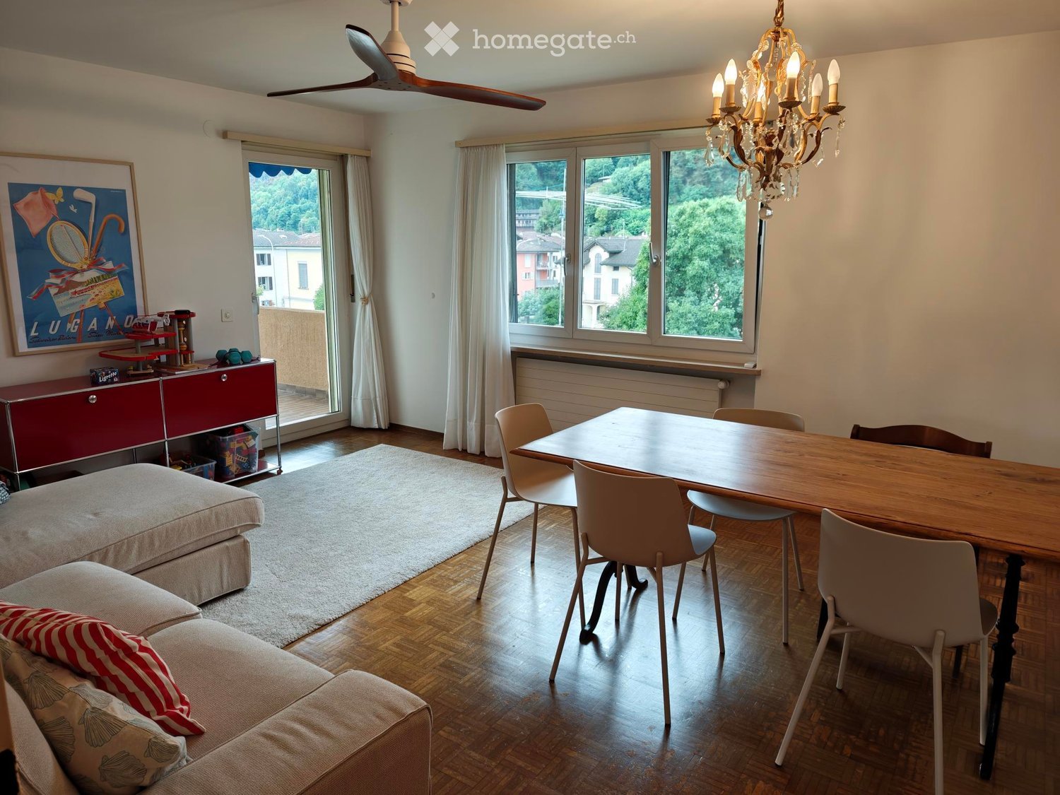 Living and dining area with a sofa, dining table, chairs, cabinet, chandelier, ceiling fan, and large windows offering a scenic view.
