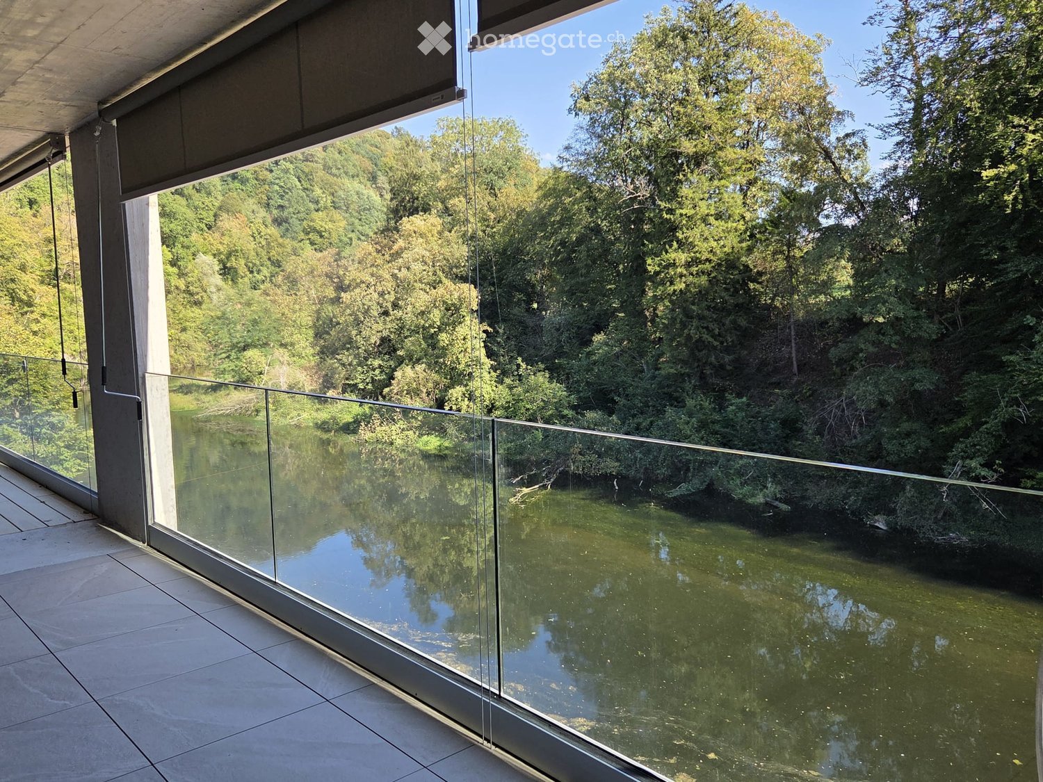 Modern balcony with large glass panels, overlooking a lush green landscape and water body.