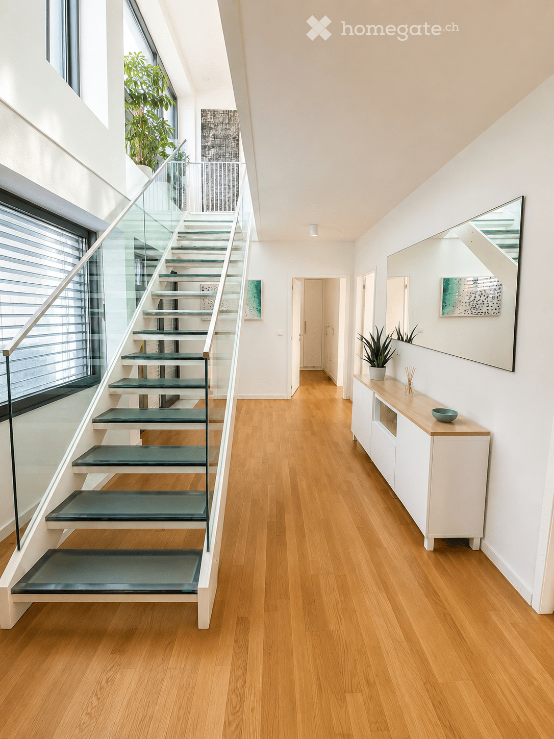 Modern hallway with wooden flooring, glass staircase with green steps, white walls, and a small console table with a plant and decorative items.