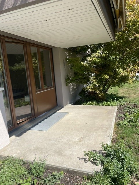 A modern balcony or terrace area with a sliding glass door, concrete floor, and surrounding greenery.