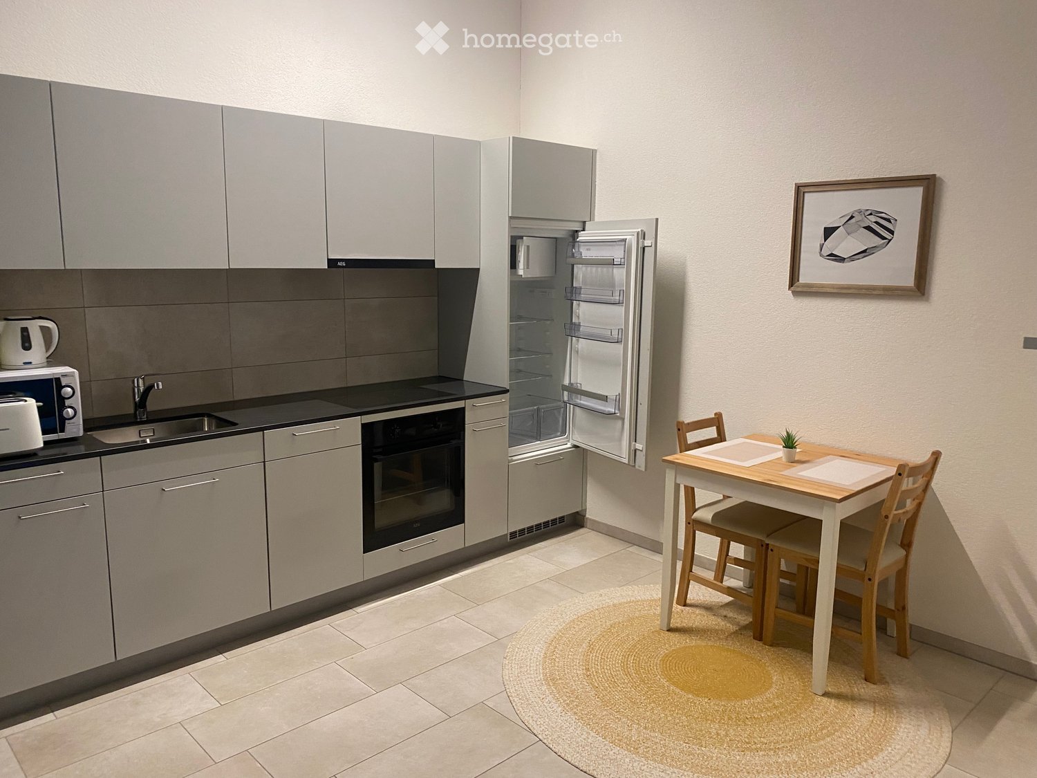 Modern kitchen with grey cabinets, stainless steel appliances, a dining area with a small table and chairs, and a round rug on the floor.