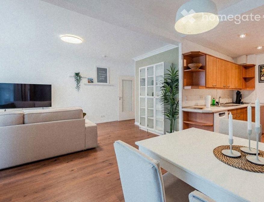 A modern, open-plan living and dining area with a light-colored sofa, a white dining table with chairs, and a small kitchen area featuring wooden cabinets and a dishwasher. The space includes a large screen, a potted plant, and decorative candles on the t