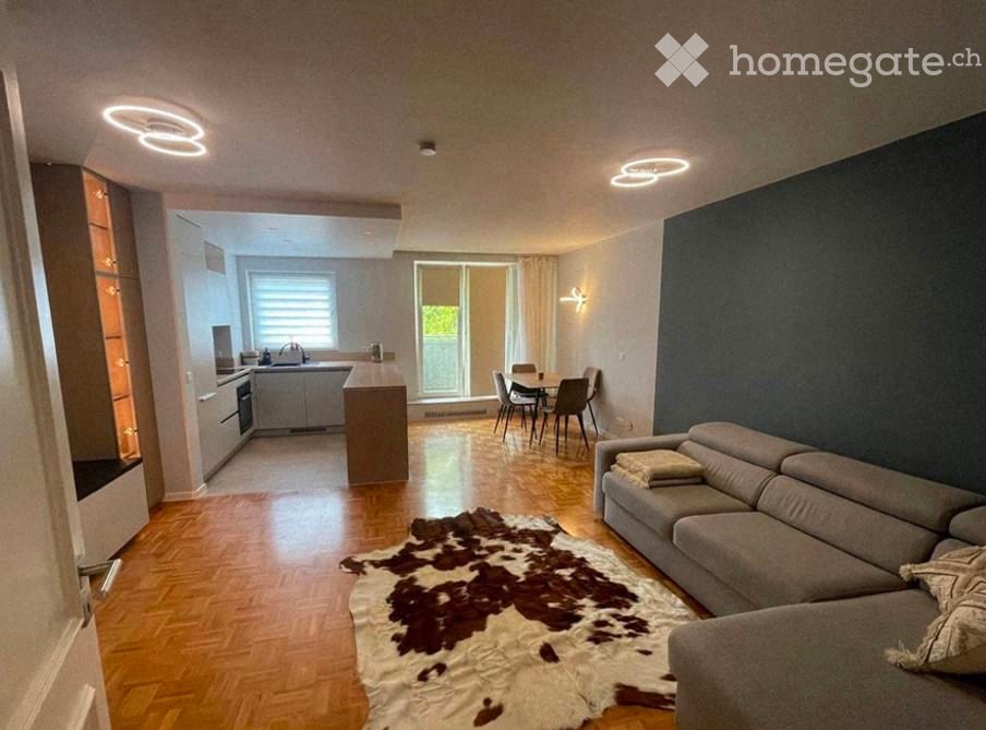 Modern open-plan living and dining area with a grey sofa, coffee table, dining table with chairs, and a small kitchen area with appliances.