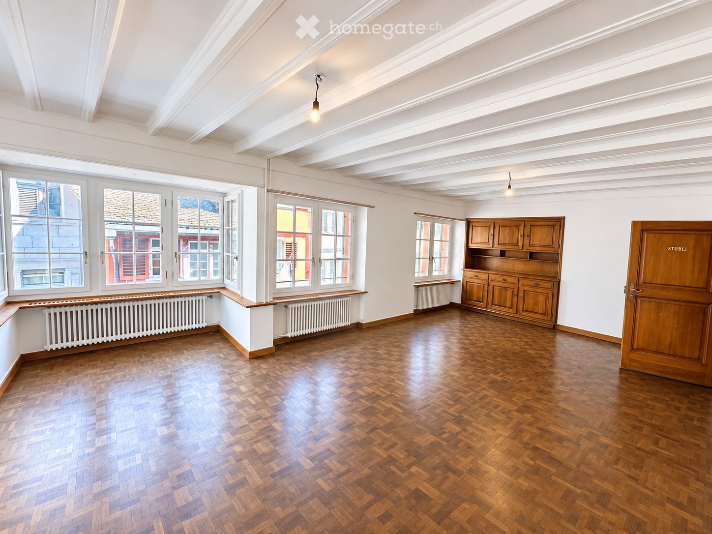 Empty room with wooden herringbone flooring, white walls, large windows, built-in wooden cabinetry, and a wooden door labeled "STUBLI".