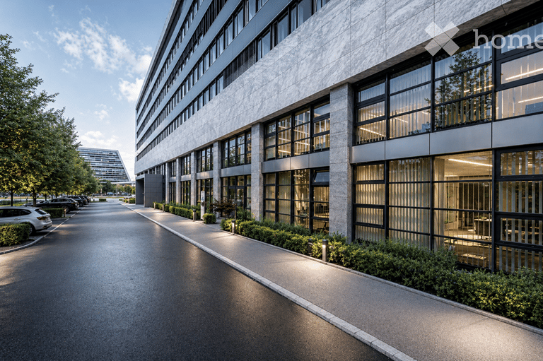Modern multi-story building with large glass windows and a clean, contemporary design. The building is surrounded by a landscaped area with trees and shrubs, and there is a paved walkway leading to the entrance. Several parked cars are visible in the fore