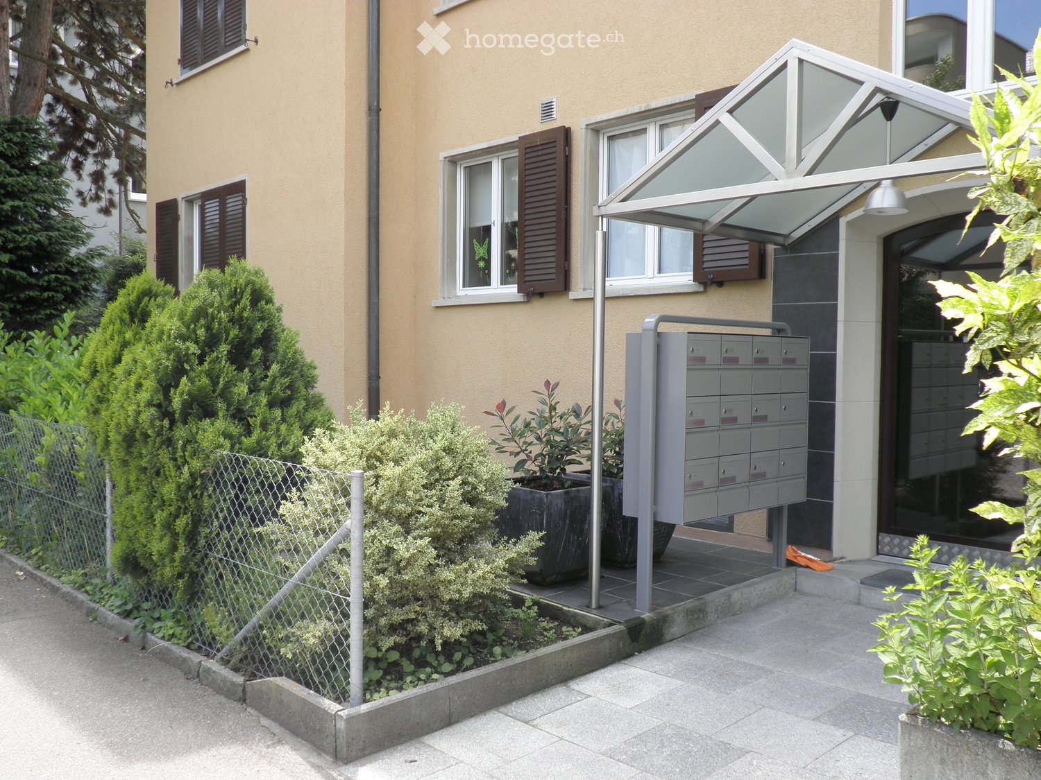 Modern building entrance with a covered porch, mailboxes, and small garden area with shrubs and potted plants.