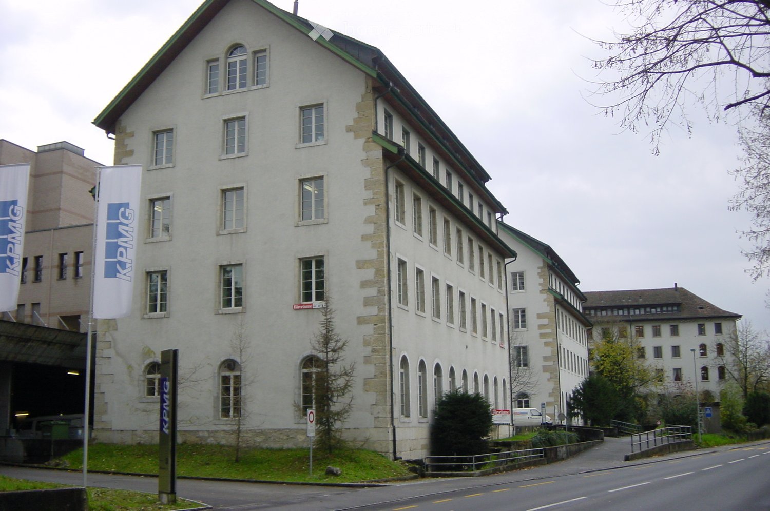 A large, multi-story building with a light-colored facade and numerous windows, featuring a green roofline and stone accents. The building has a modern design with a mix of rectangular and arched windows, and is surrounded by a paved area with a small gar