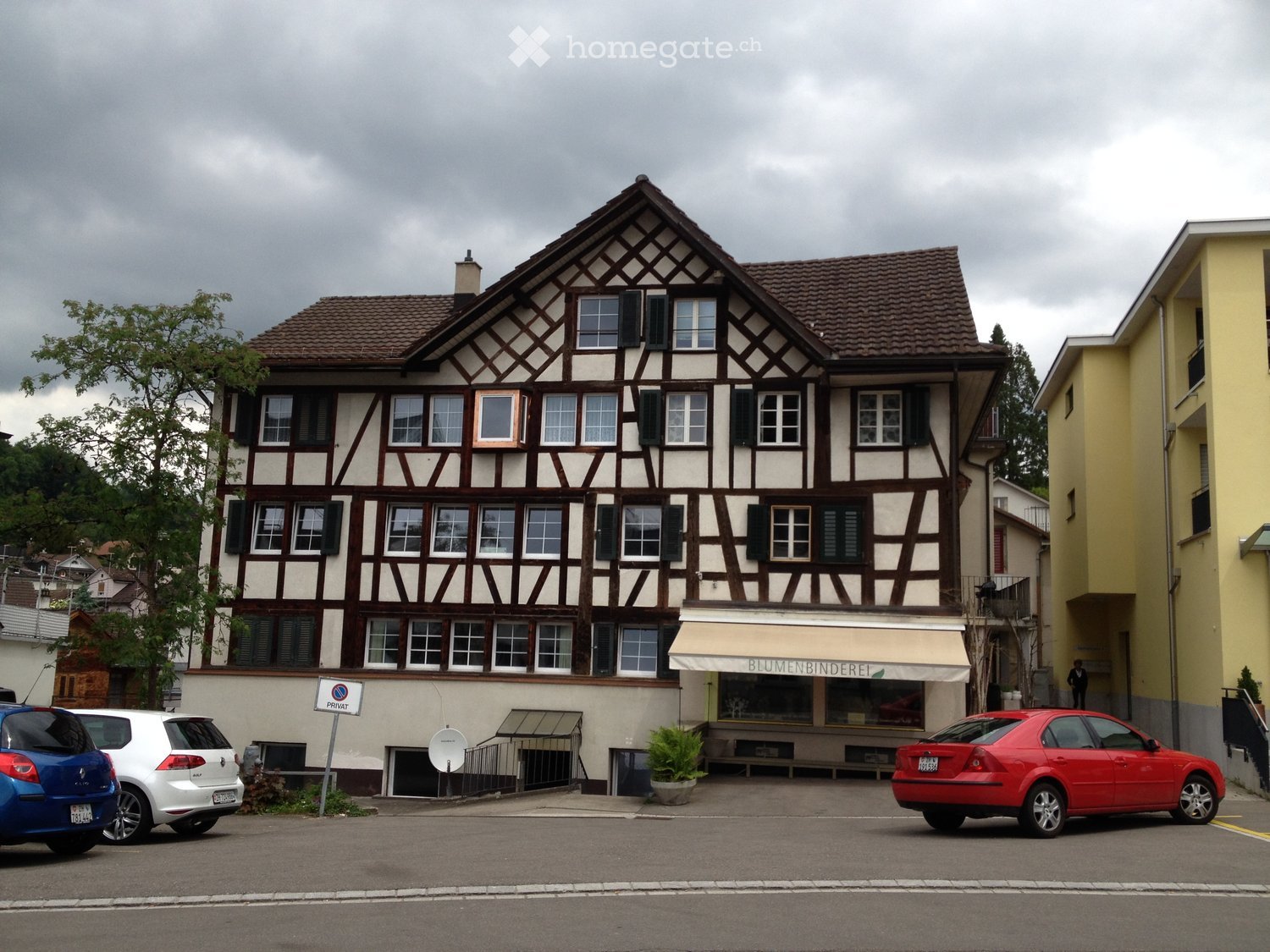 2 story building, beige paint, brown roof, windows with shutters, ground floor awning, parking area