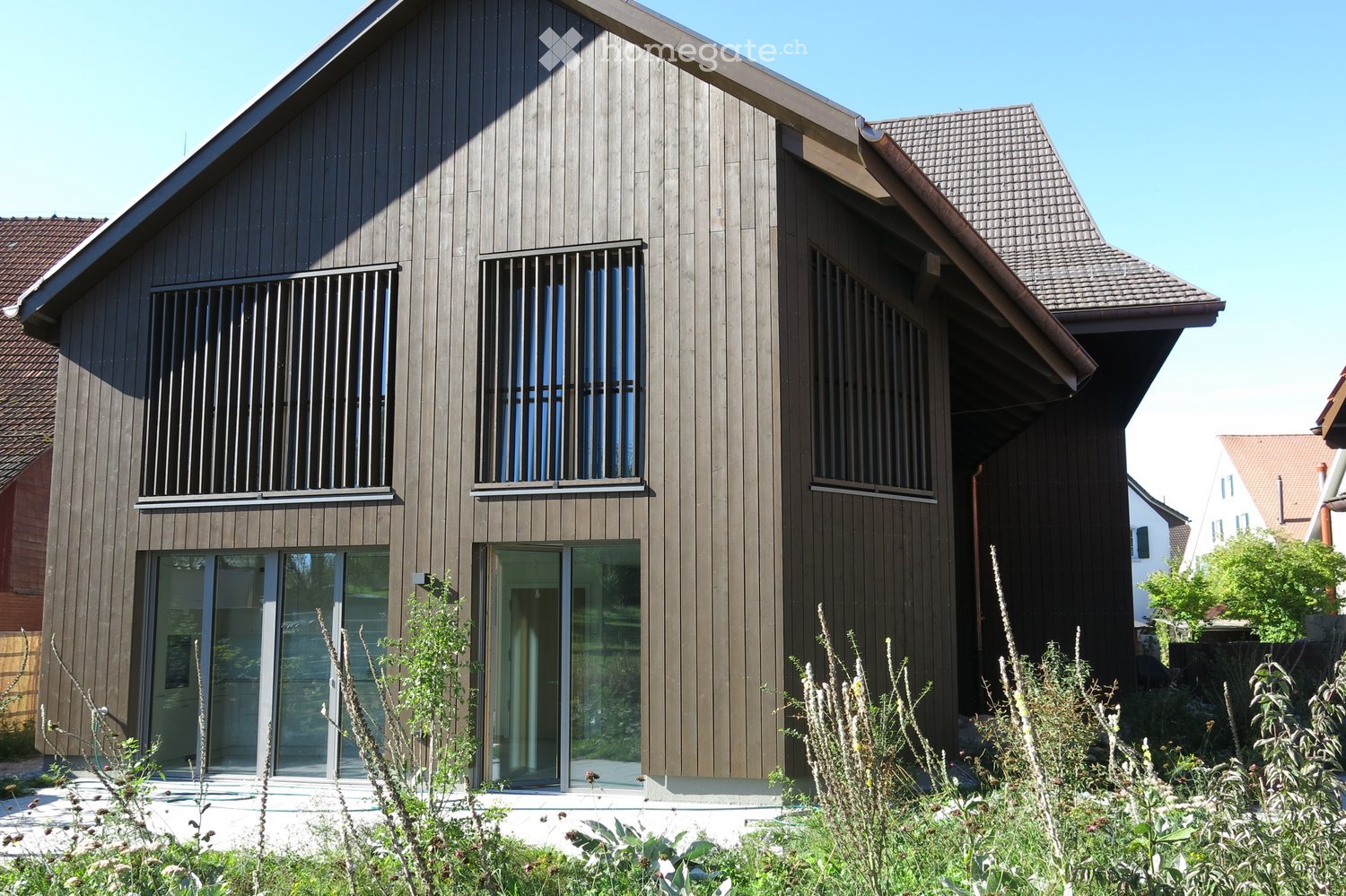 Modern two-story house with dark wooden siding, large windows, and a sloped roof. Features include a covered entrance and surrounding greenery.