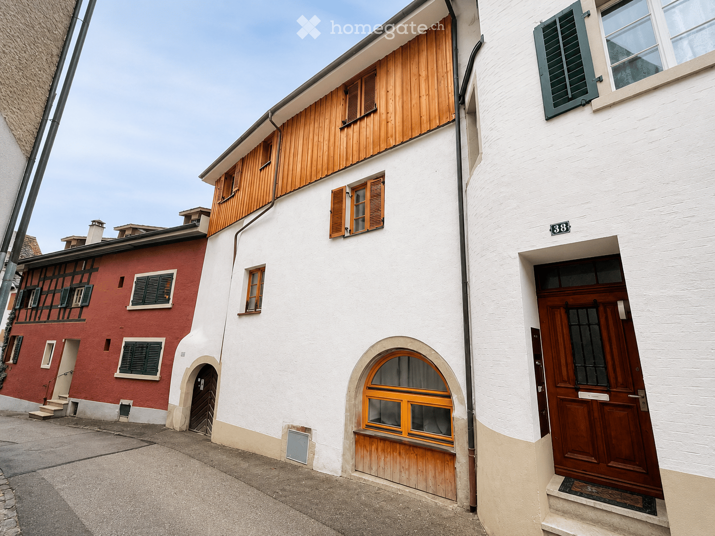A narrow alleyway with a two-story building featuring white walls, wooden shutters, and a wooden door. The building has a small arched entrance and a wooden garage door. The adjacent building is red with green shutters and a small staircase leading to an