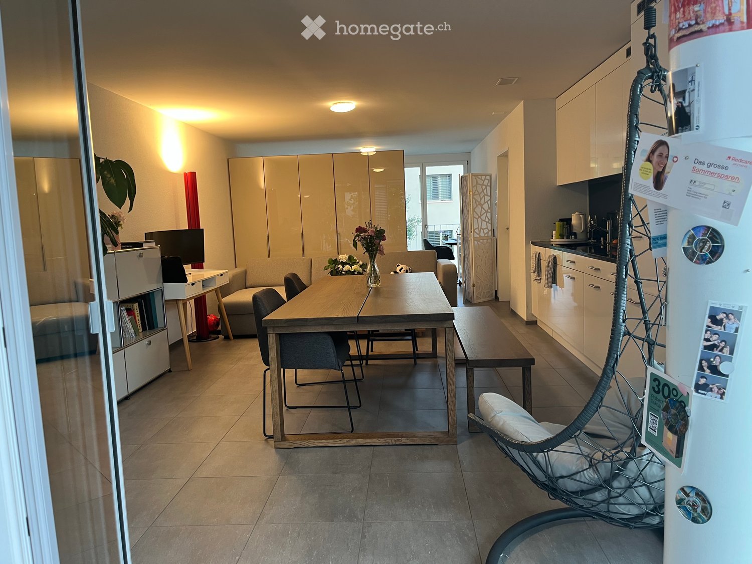 A modern, well-lit living and dining area with a sofa, dining table, chairs, kitchen cabinets, and a refrigerator. The space features a wicker hanging chair, a small bench, and a flower vase on the table.