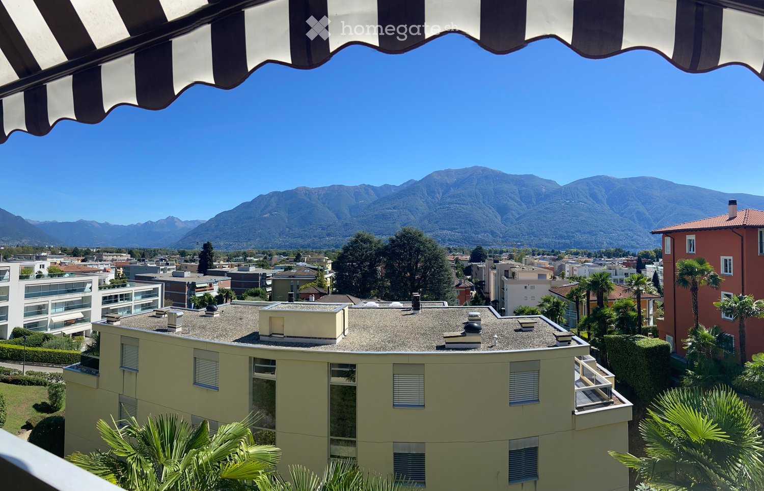 Balcony with striped awning, overlooking a cityscape with buildings, trees, and distant mountains under a clear blue sky.