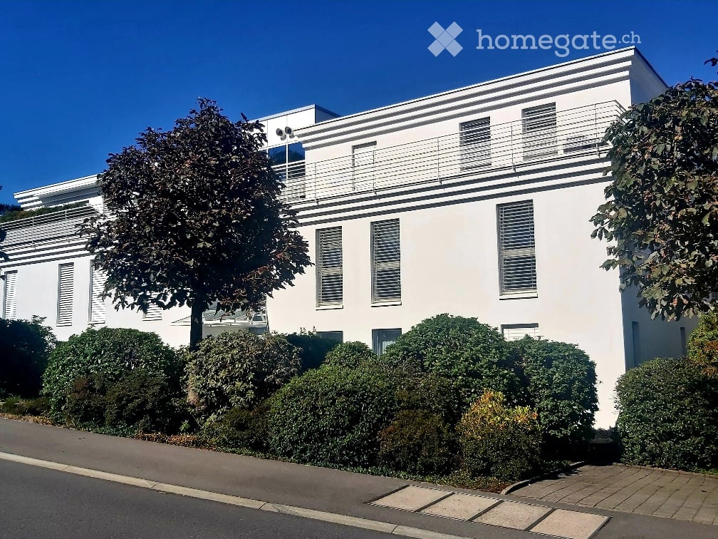 Modern two-story white building with large windows, balconies, and trimmed hedges in front.