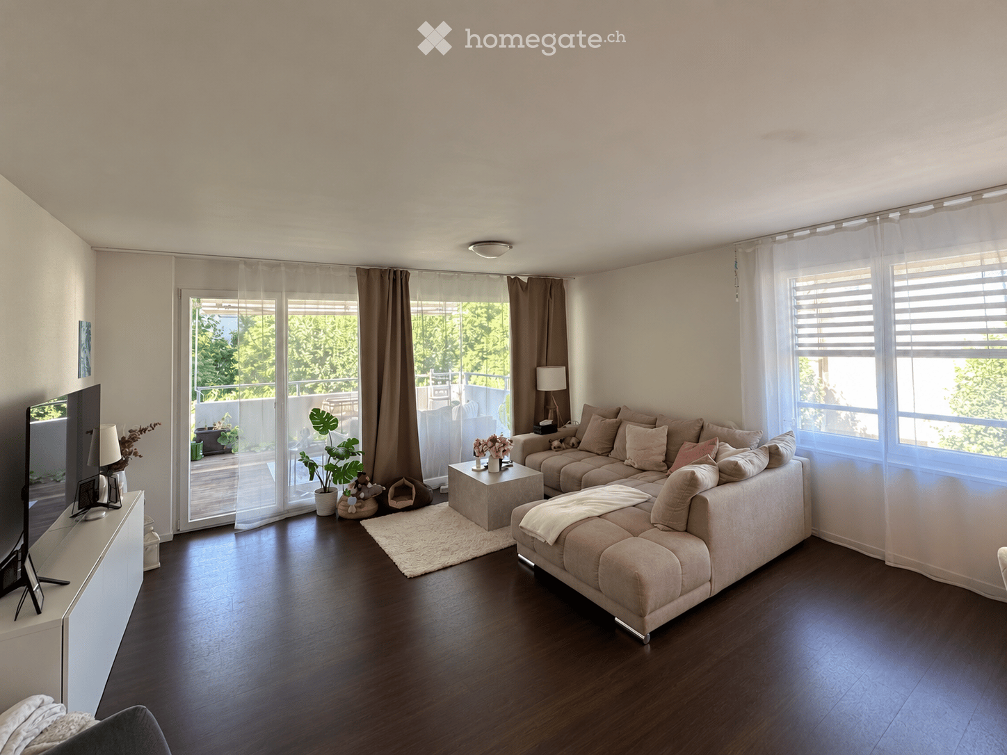 Modern living room with hardwood flooring, large windows, and a balcony. Features a beige sectional sofa, coffee table, TV, and decorative plants.