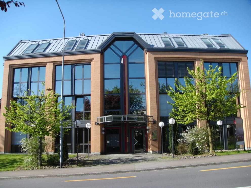 Modern multi-story building with large glass windows and a sleek, contemporary design. The entrance features a covered walkway with a glass canopy, flanked by trees and street lamps.